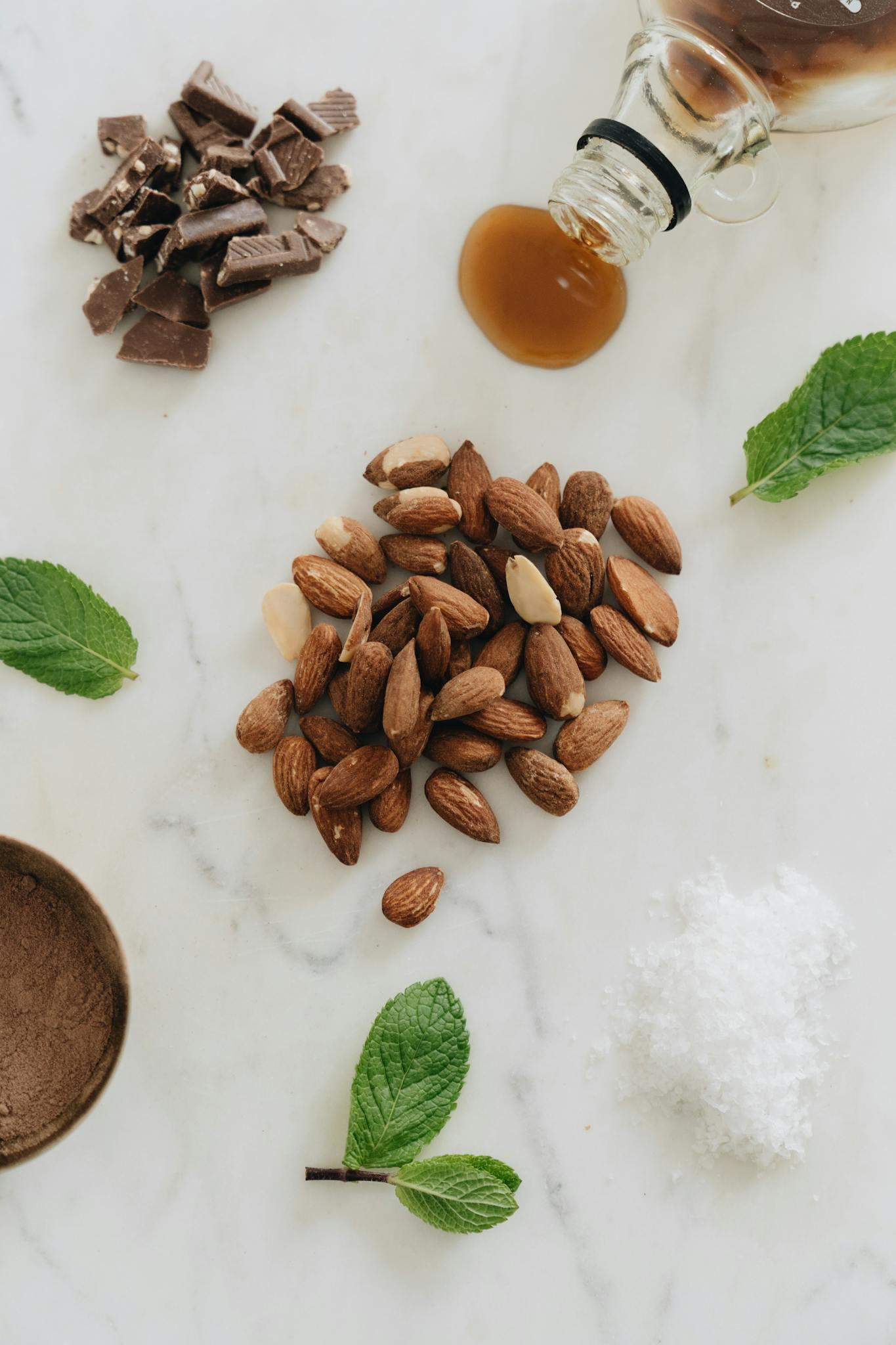 A flat lay of almonds, chocolate, mint, and maple syrup on marble.
