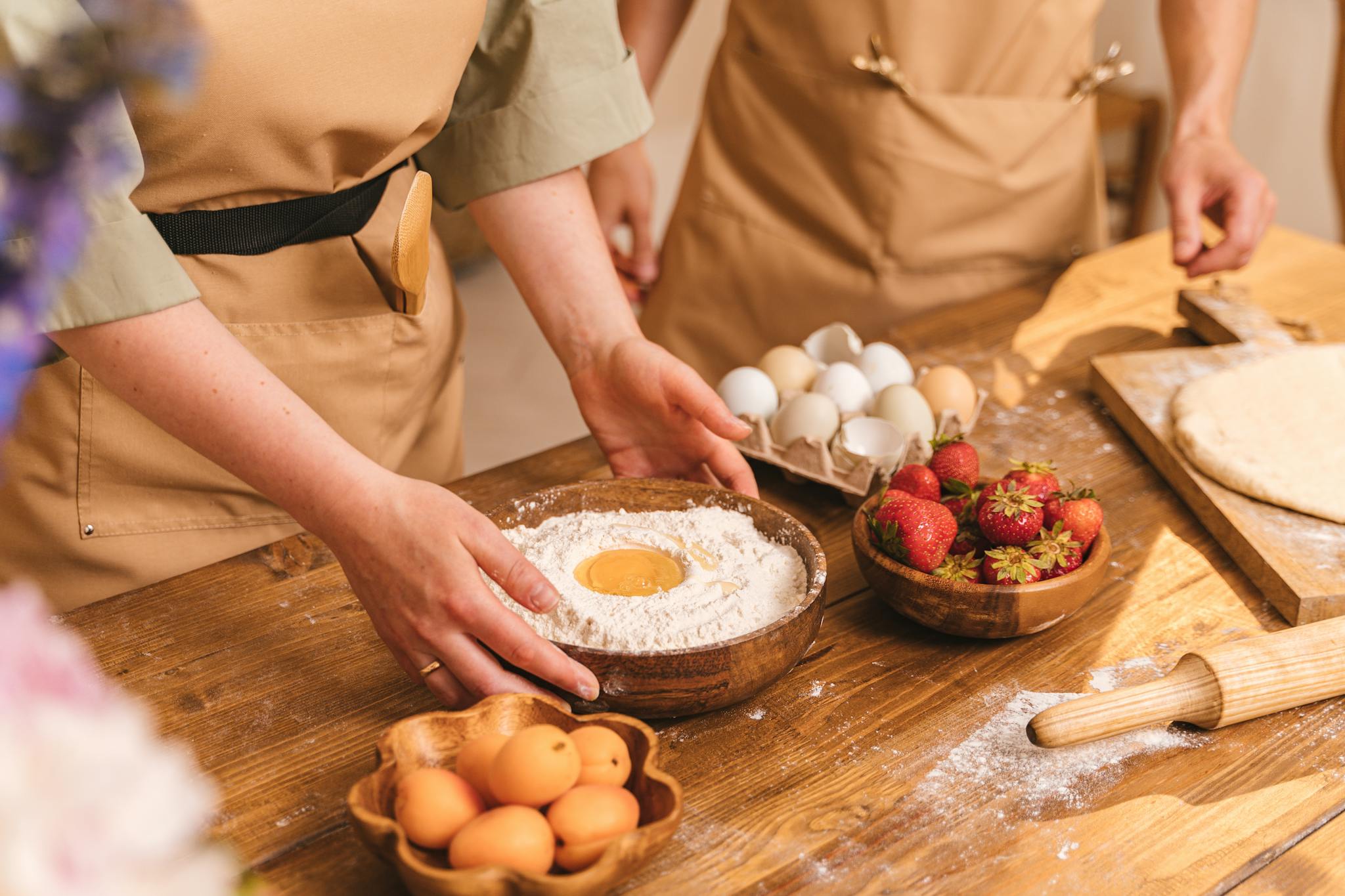 Adults preparing dough with eggs, flour, and strawberries on a wooden counter.