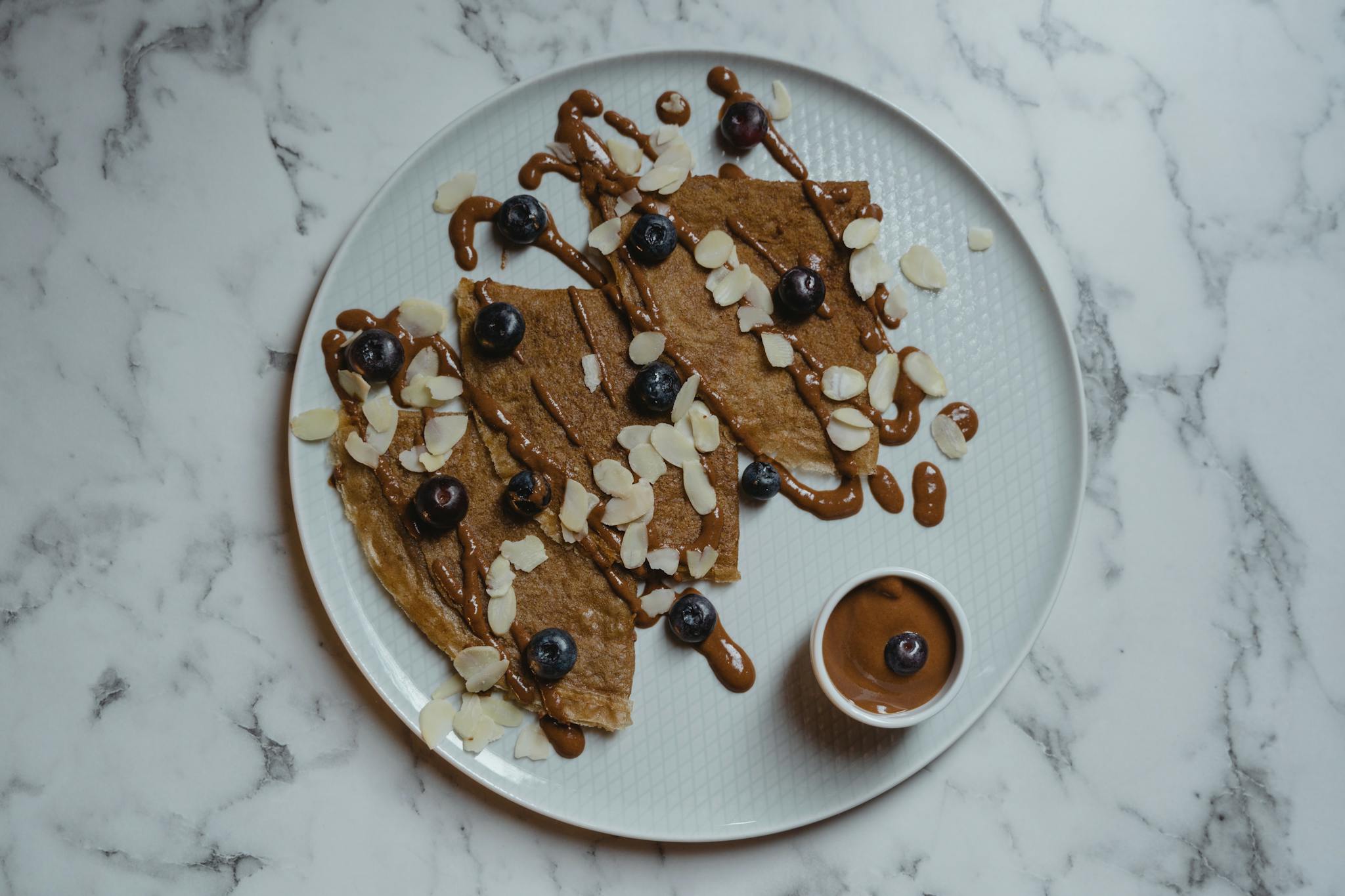 Delicious crepes topped with blueberries and almond slices, drizzled with chocolate sauce on a marble surface.
